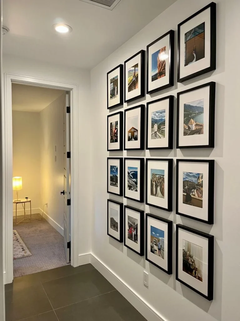 Modern Grid Hallway Gallery Wall A neat grid layout of sixteen matching black framed photos with white matting displayed in a modern white hallway, leading to an open doorway with a lit lamp and side table beyond.
