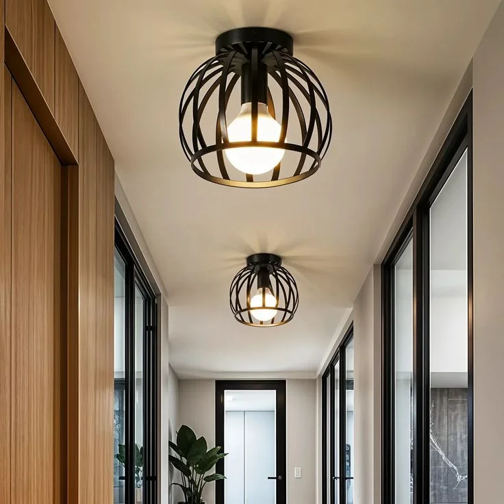 Two modern pendant ceiling lights with black metal cage shades illuminating a minimalist hallway with wooden wall panels and glass doors.