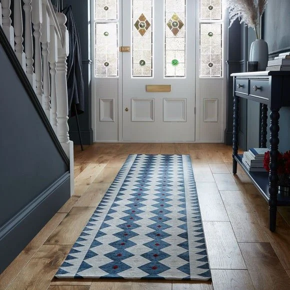 A long hallway runner rug featuring a blue and white geometric diamond pattern with small red accents, laid out on light oak wooden flooring. The hallway includes a dark blue accent wall, a white wooden staircase banister, and a black console table leading toward a white front door with stained glass panels.