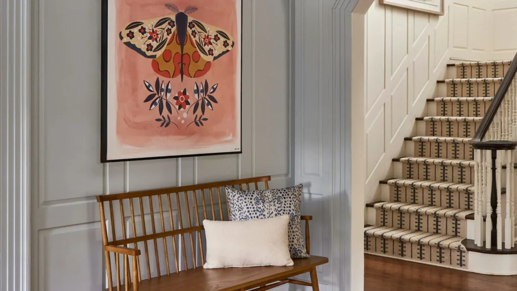 A cozy hallway nook featuring a wooden spindle bench decorated with a blue-spotted throw pillow and a small cream cushion. Above the bench hangs a large framed art piece depicting a stylized moth with floral wings on a dusty pink background. The light grey paneled walls lead to an adjacent staircase runner rug with a cream and black striped geometric pattern installed over wooden steps.