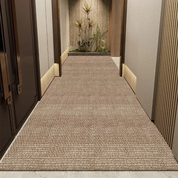 A wide, textured light brown sisal style carpet runner stretching down a luxury modern corridor. The hallway features neutral paneled walls with warm recessed baseboard lighting, dark double doors on the left, and an indoor plant alcove at the far end.