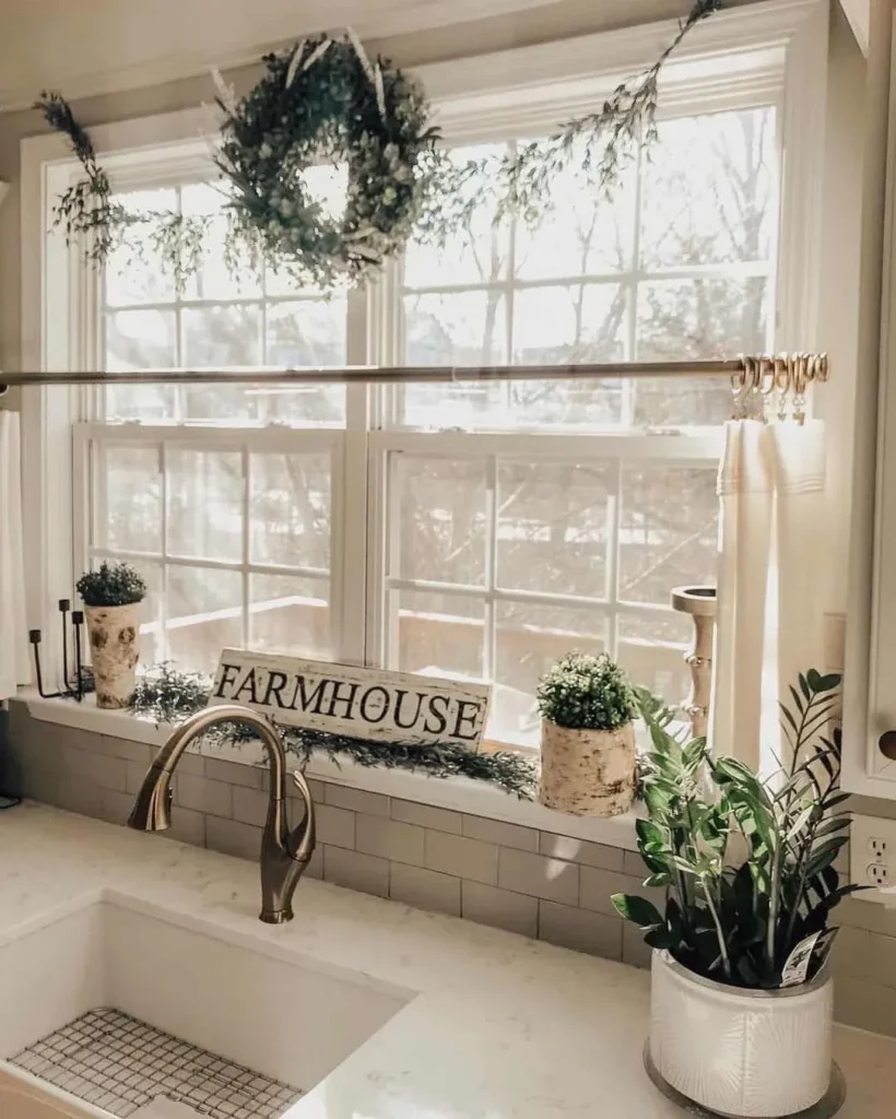 A bright, modern farmhouse kitchen window styled with a green garland and a central wreath across the top. The windowsill features a rustic wooden "FARMHOUSE" sign alongside small potted plants in textured birch bark containers. Below, a brass gooseneck faucet sits by a white sink, with a vibrant potted ZZ plant resting on the quartz countertop.