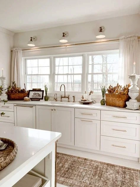 A bright white kitchen featuring a large triple window framed by soft white curtains on a continuous brass rod and illuminated by three overhead sconces. The white countertop holds a sink with a brass gooseneck faucet, flanked by woven storage baskets filled with wooden utensils. A patterned brown vintage runner rug coordinates with the warm metallic details to complete the cozy, neutral space.
