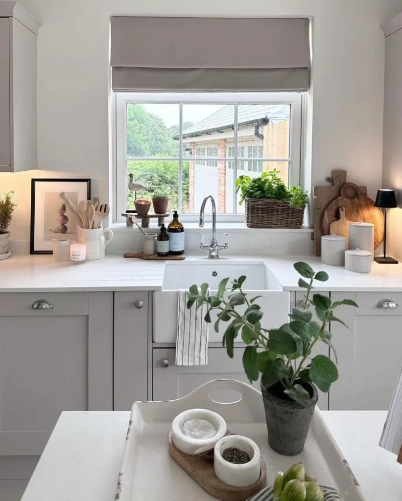 A well-styled kitchen sink area featuring a white ceramic apron sink with a chrome gooseneck faucet. The windowsill holds a wicker basket with herbs and other plants, while a beige Roman blind is pulled down above.