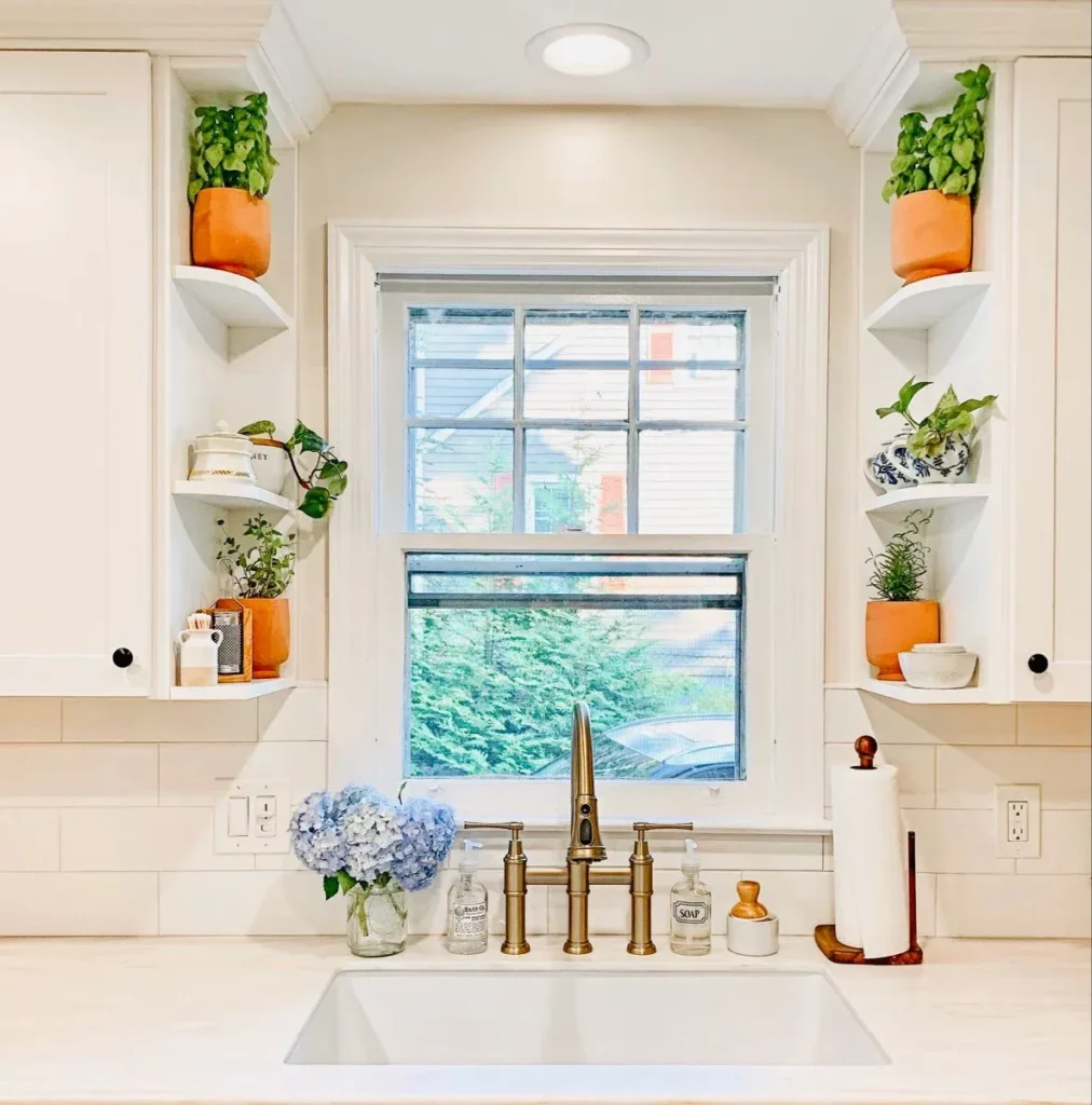 A symmetrical white kitchen sink area with a brass bridge faucet below a central window. Built-in corner display shelves flank the window, holding green houseplants and herbs in terracotta pots. A white subway tile backsplash frames the area, while a glass vase filled with blue hydrangeas and a wooden paper towel holder rest on the counter.
