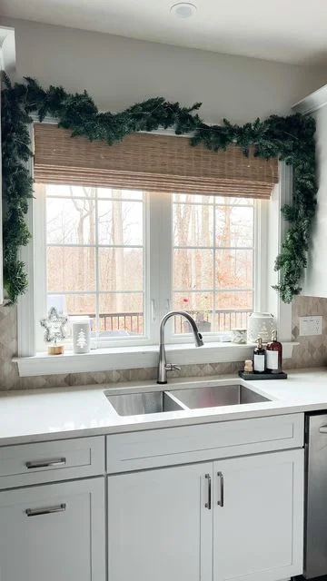A bright kitchen window area styled for the holidays with a thick green pine garland draped around the white window frame. A woven bamboo Roman shade sits above a double-bowl stainless steel sink equipped with a brushed nickel gooseneck faucet. Small festive winter decorations, including a star and a tree silhouette, rest on the windowsill against a patterned backsplash tile.