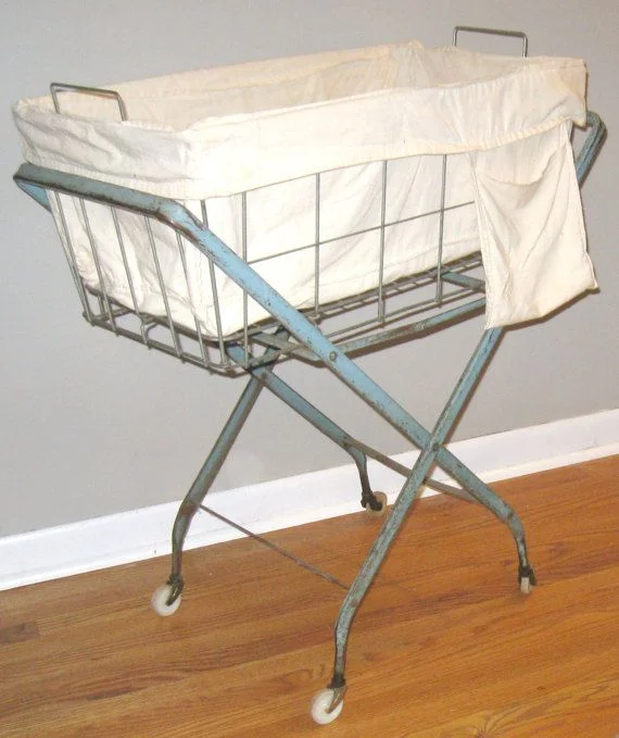 Vintage light blue metal wire laundry cart on wheels with an off-white canvas fabric liner and side pocket, set on a hardwood floor.