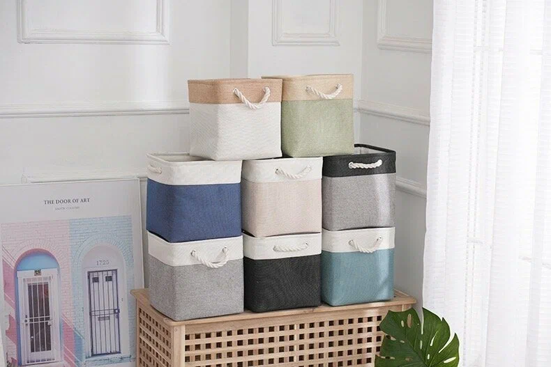 Stack of rectangular two-tone fabric laundry storage bins in various colors with thick white rope handles set on a wooden cabinet.