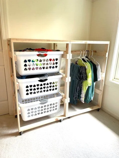 Light wood DIY laundry storage unit on caster wheels. The left side holds three stacked white plastic laundry baskets sliding on wooden tracks. The right side features a hanging rod displaying several shirts on hangers.