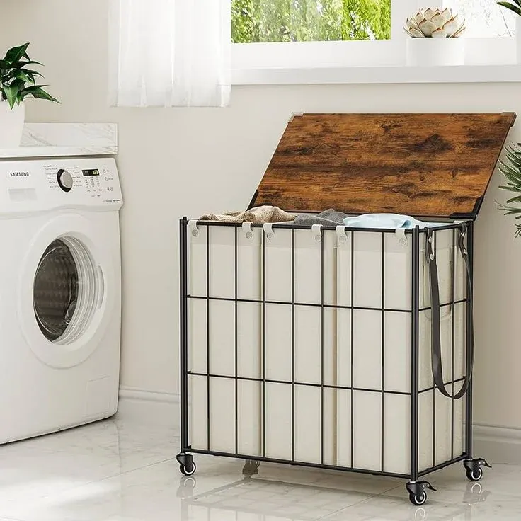 An industrial-style rolling laundry sorter with a black wire frame, three removable beige fabric bags, and a hinged rustic brown wooden lid that is open, revealing clothes inside. The cart is on wheels in a white laundry room next to a washing machine.