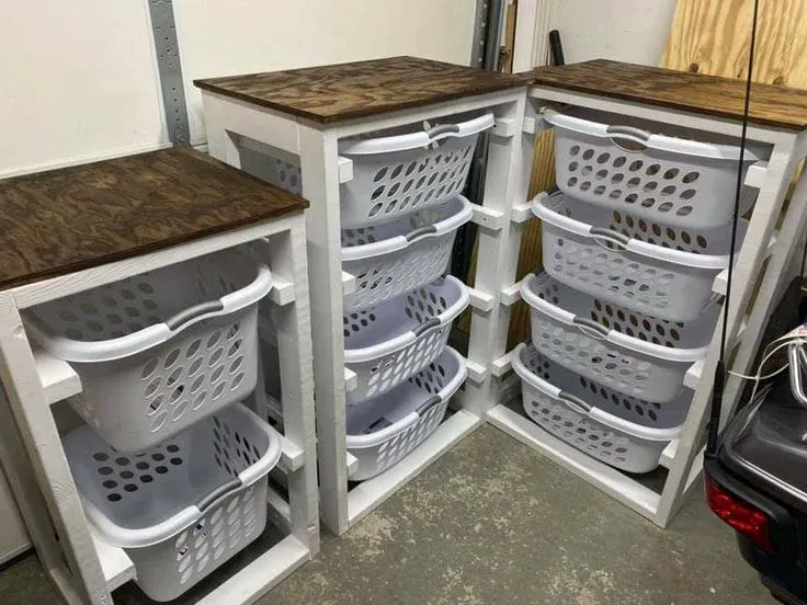 A modular DIY storage setup featuring three painted white wooden frames with stained dark wood countertops. The left tower holds two white plastic laundry baskets, while the middle and right towers hold four baskets each, sliding out like drawers.