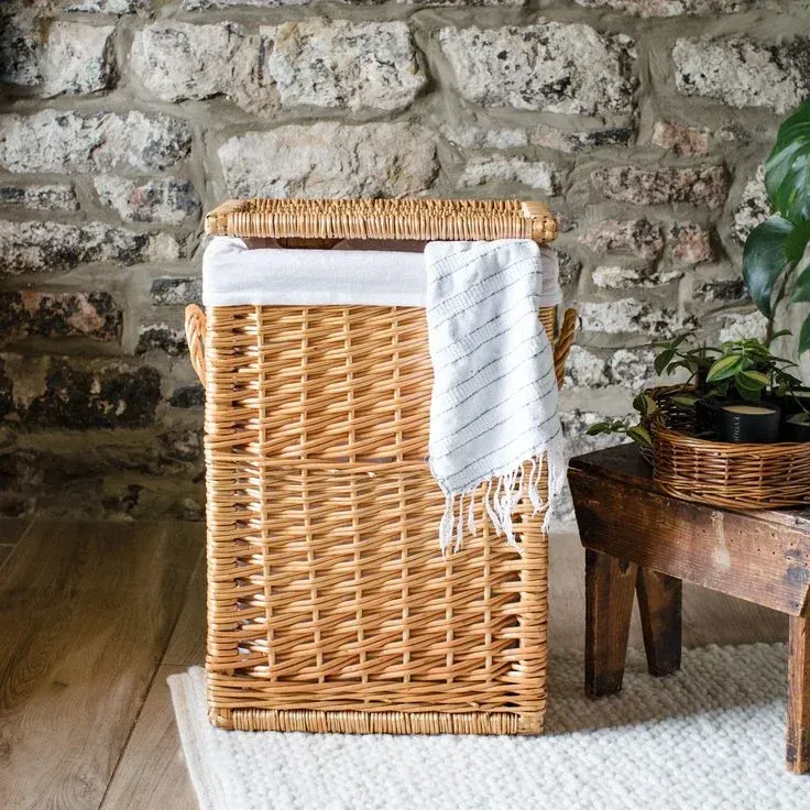 Rectangular woven wicker laundry basket with a lid, removable white fabric liner, and side handles, placed in front of a rustic stone wall next to a wooden stool.
