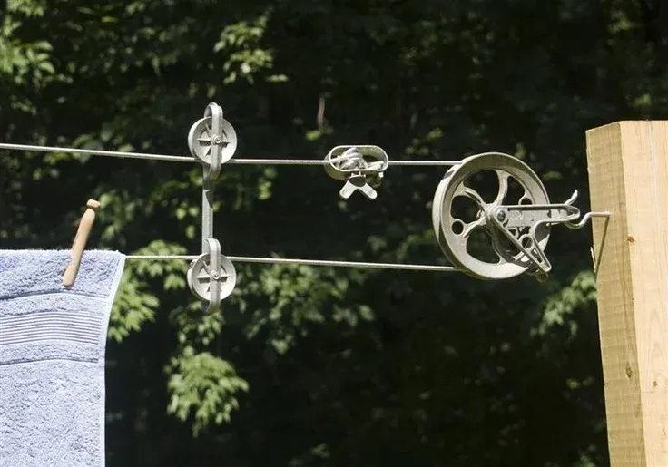 Close-up of an outdoor pulley clothesline mechanism mounted to a wooden post, featuring metal wheels, line tighteners, line separators, and a blue towel secured to the wire with a wooden clothespin against a lush green foliage backdrop.