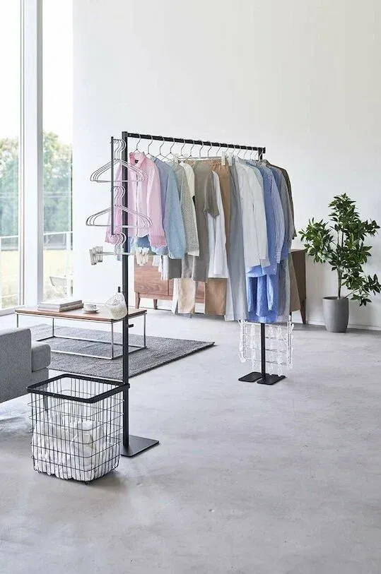 A sleek, black freestanding metal garment rack loaded with hanging shirts and trousers in a modern, brightly lit living space, featuring side hooks for extra hangers and an attached wire laundry basket near the base.