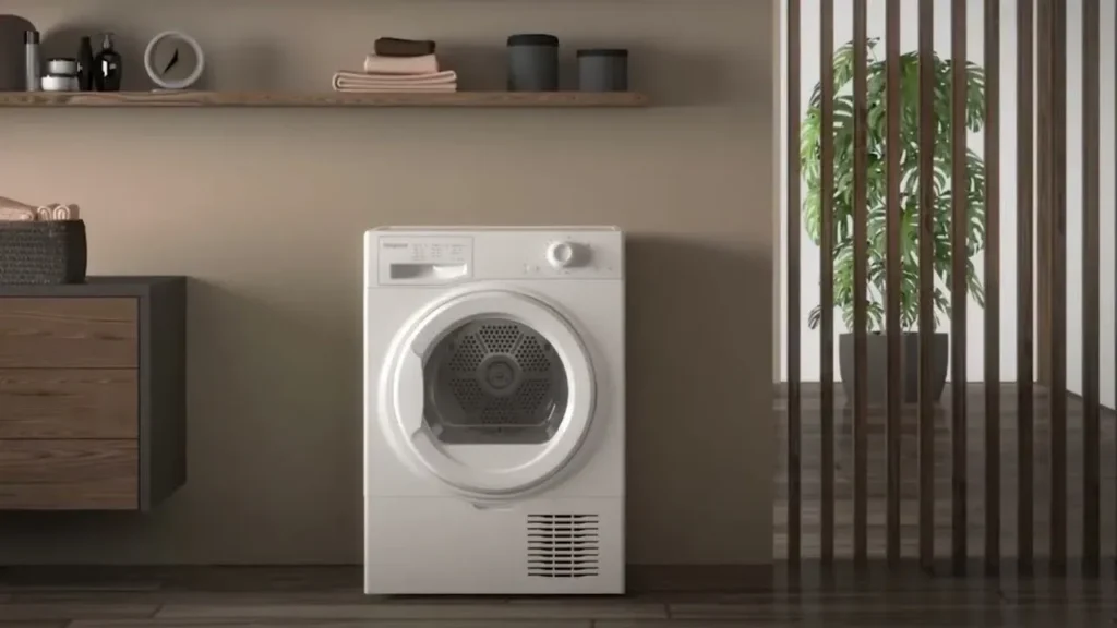 A white freestanding front-loading tumble dryer positioned in a modern, minimalist laundry space with dark wood flooring, beige walls, floating wooden shelves, and a slatted wooden room divider revealing a green potted plant in the background.