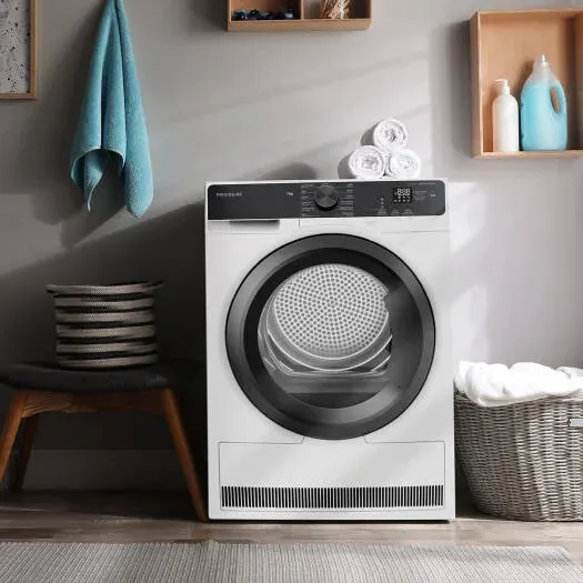 A white Frigidaire front-loading clothes dryer with a dark grey control panel, set against light grey walls with floating wooden shelving containing detergent bottles, rolled towels above, a blue hanging hand towel, and a woven laundry basket nearby.