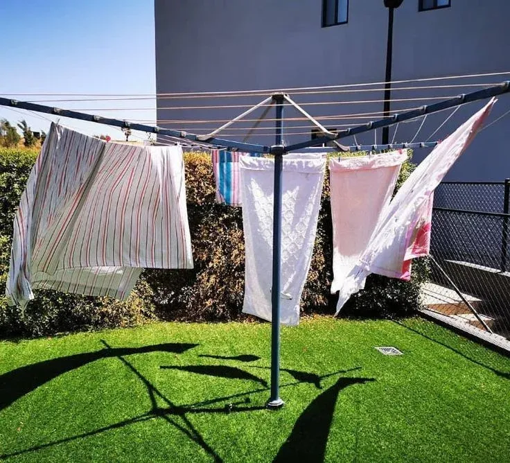 An outdoor umbrella-style rotary clothes airer set up on green lawn grass, with various laundry items including bed sheets and white towels hanging to dry in the sunshine.