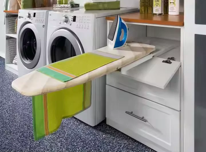 A space-saving laundry room featuring a pull-out ironing board extended from a white built-in cabinet. A blue and white clothes iron rests on the board, and a bright green towel hangs over the side. In the background, a white front-loading washer and dryer set sits alongside the cabinetry.