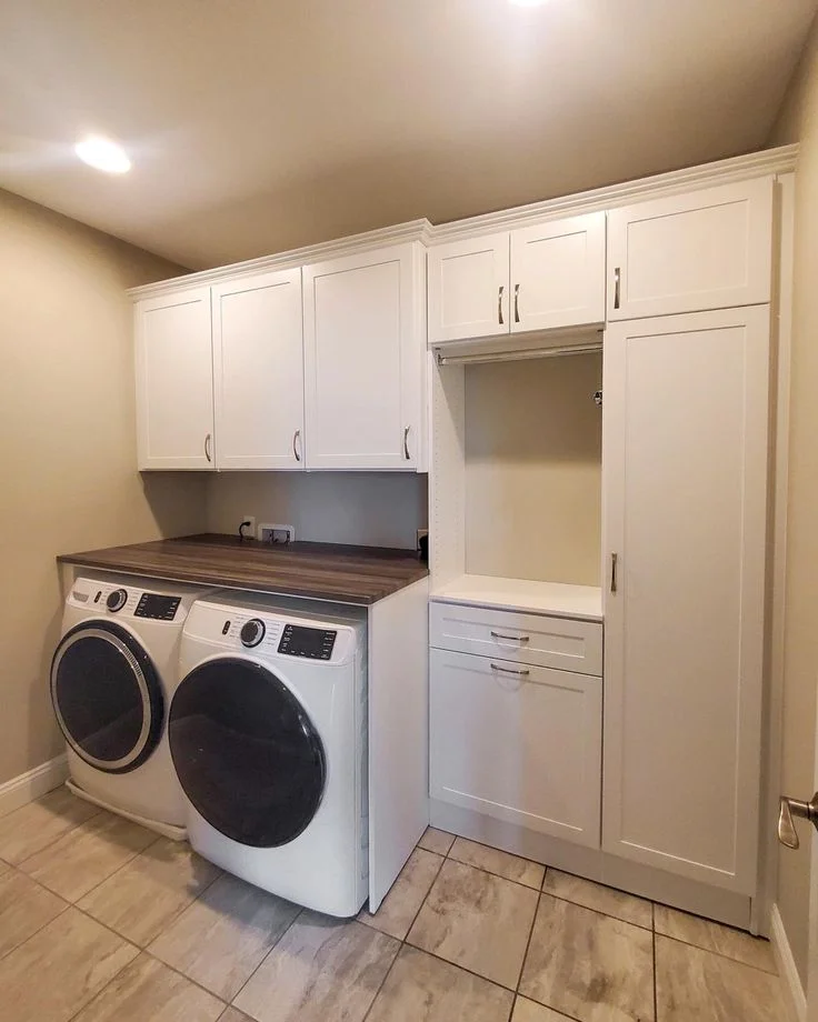 A bright laundry room featuring white front-loading washing and drying machines under a dark wood countertop. Extensive custom white cabinetry includes upper cabinets above the machines, a tall pantry cabinet, lower drawers, and an open nook with a hanging rod.