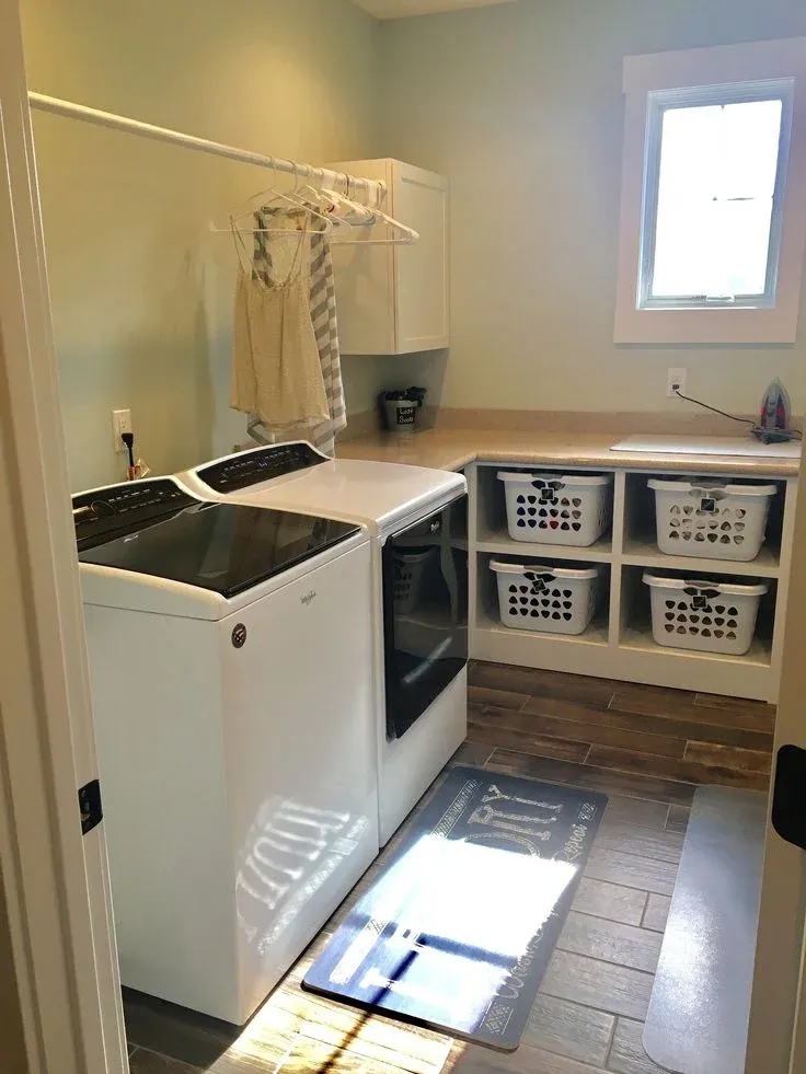 A sunlit, spacious laundry room featuring a white top-load washer and front-load dryer set alongside an L-shaped counter. Beneath the countertop are built-in open cubbies holding four white plastic laundry baskets. A clothing rod with hung shirts extends above the washer, and light green walls frame a small window.