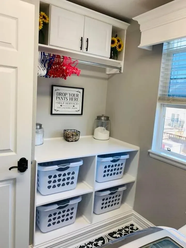 Organized white laundry room station featuring four lower cubbies with light blue plastic laundry baskets, a white counter for folding, a framed funny sign reading "Drop Your Pants Here," a clothes hanging rod with colorful hangers, and upper cabinets flanked by faux sunflowers.