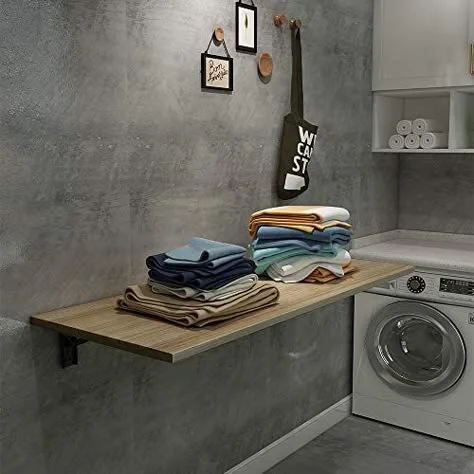 A space-saving, wall-mounted light wood drop-leaf folding table installed against a dark concrete-style wall with stacks of folded colorful towels on top, located next to a front-load washing machine.