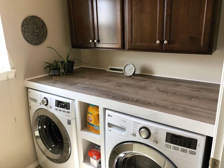 A modern laundry room featuring a white LG front-loading washer and dryer built under a dark wood-grain folding counter. A small white shelf unit sits between the appliances holding detergent and dryer sheets. Above the counter are dark wooden upper cabinets, potted aloe and succulent plants, a small round clock, and a sun-face wall plaque on the left wall.