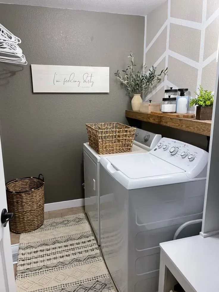 Cozy laundry room setup featuring top-load washing machine and dryer, a dark wood floating shelf with decorative jars and plants, a white wall sign reading "I'm feeling dirty", a geometric patterned accent wall, woven storage baskets, and a boho runner rug.