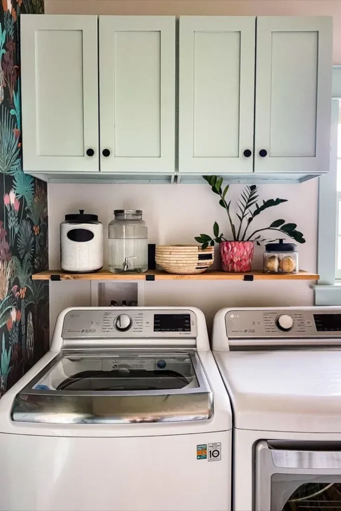 Bright laundry room featuring white LG top-load washer and dryer units, pale mint green shaker upper cabinets, a light wood floating shelf styled with glass jars and potted plants, and a vibrant botanical wallpaper accent wall.