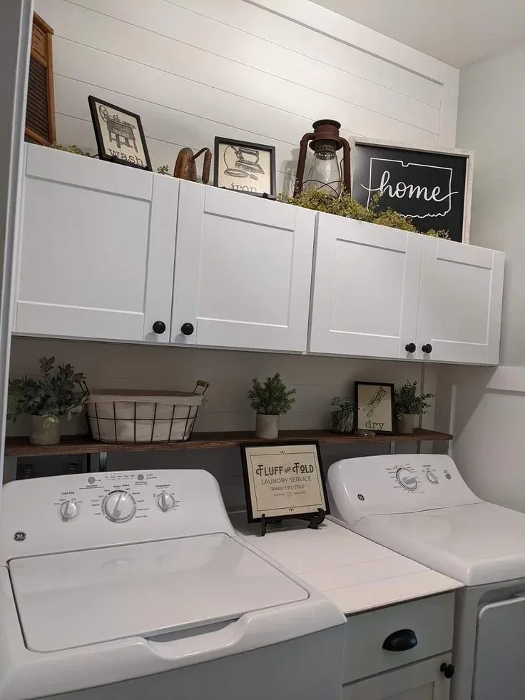 Rustic farmhouse laundry room featuring white GE top-load washer and dryer units, white shiplap accent wall, white shaker upper cabinetry styled with vintage decor like a lantern and framed signs, and a narrow wood shelf holding small potted greenery and storage baskets.