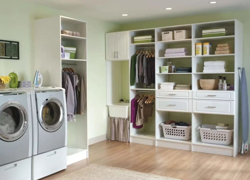 Spacious laundry room featuring white custom built-in organizer shelving with hanging rods, drawers, laundry baskets, front-load washer and dryer, and an ironing board setup.