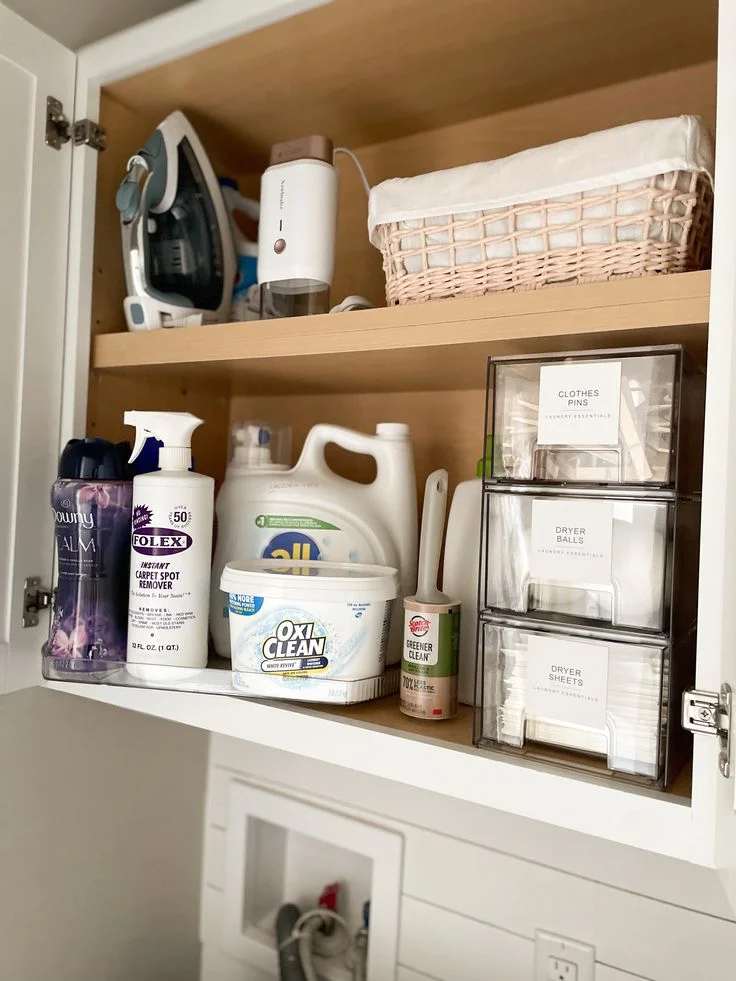 Open white laundry cabinet neatly organized with laundry detergents, stain removers, an iron, a fabric steamer, and stacked clear acrylic drawers labeled for clothespins, dryer balls, and dryer sheets.