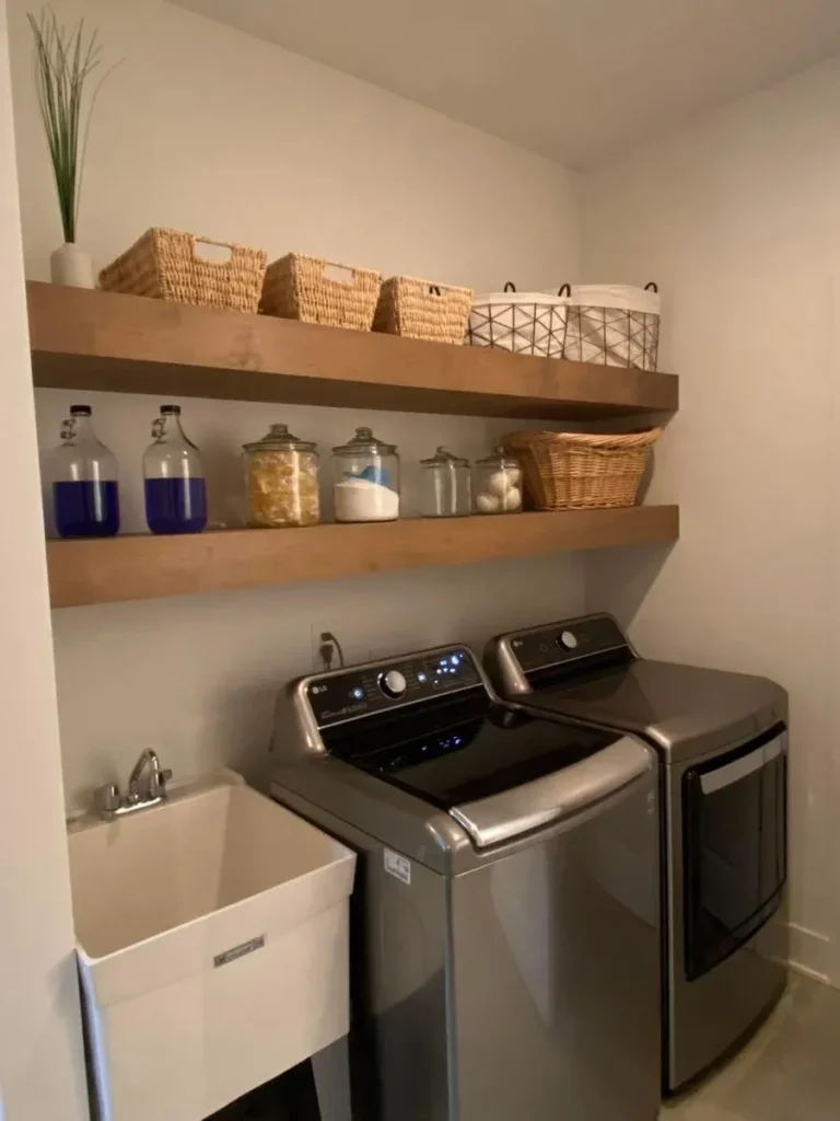 A organized laundry room featuring thick floating wooden shelves lined with woven storage baskets and glass detergent dispensers, mounted above metallic grey LG washer and dryer units and a white floor-standing utility tub sink.