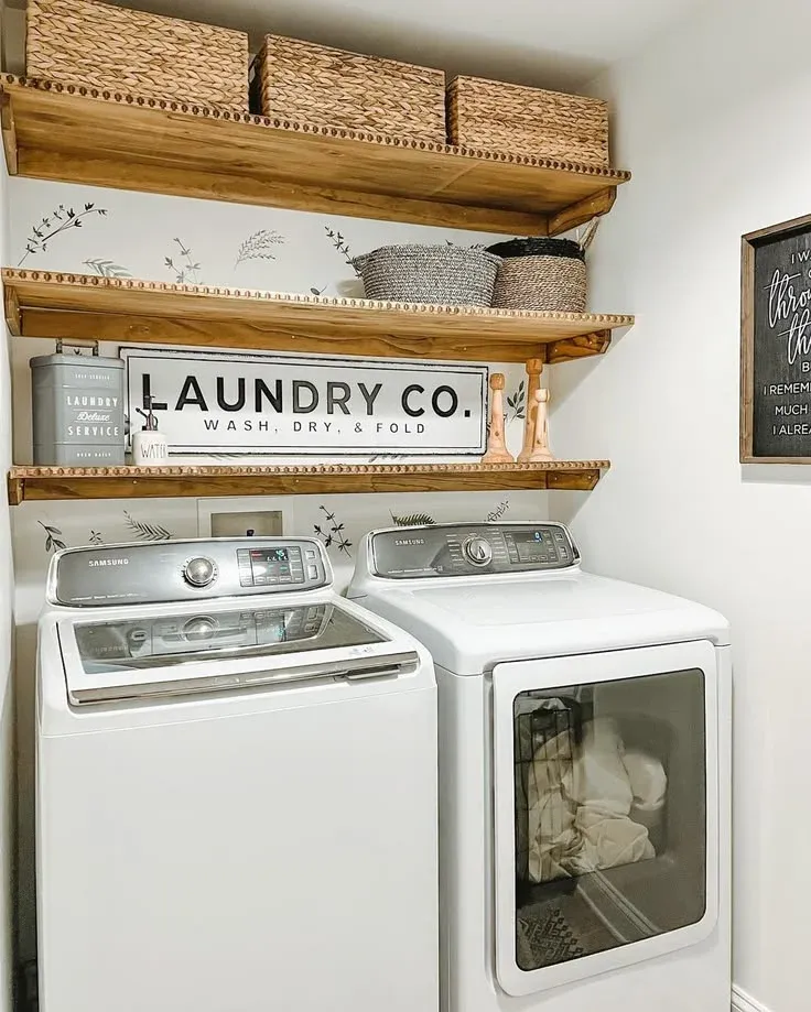A white laundry nook featuring three wooden floating shelves with beaded trim, lined with woven seagrass storage bins and decorative accents, positioned above a framed "LAUNDRY CO." sign and top-loading white Samsung washing machine and dryer units set against leaf-patterned wallpaper.
