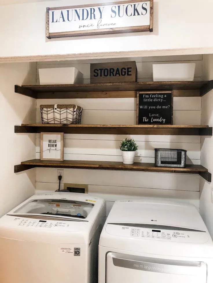 A laundry nook featuring white shiplap walls, dark wooden open shelves with storage bins, potted greenery, and small framed signs, topped by a wall sign reading "LAUNDRY SUCKS since forever" above top-loading white LG washer and dryer units.