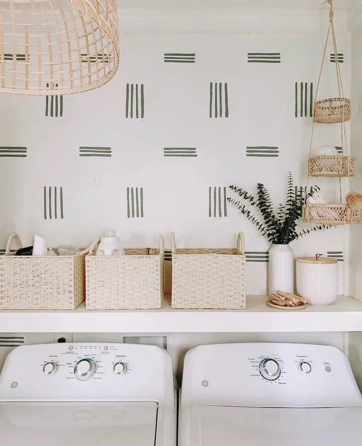 A brightly lit laundry room featuring white GE washing and drying units below a shelf organized with woven baskets, a vase of eucalyptus, and a hanging macrame organizer, set against a white wall decorated with a minimalist black striped geometric pattern.