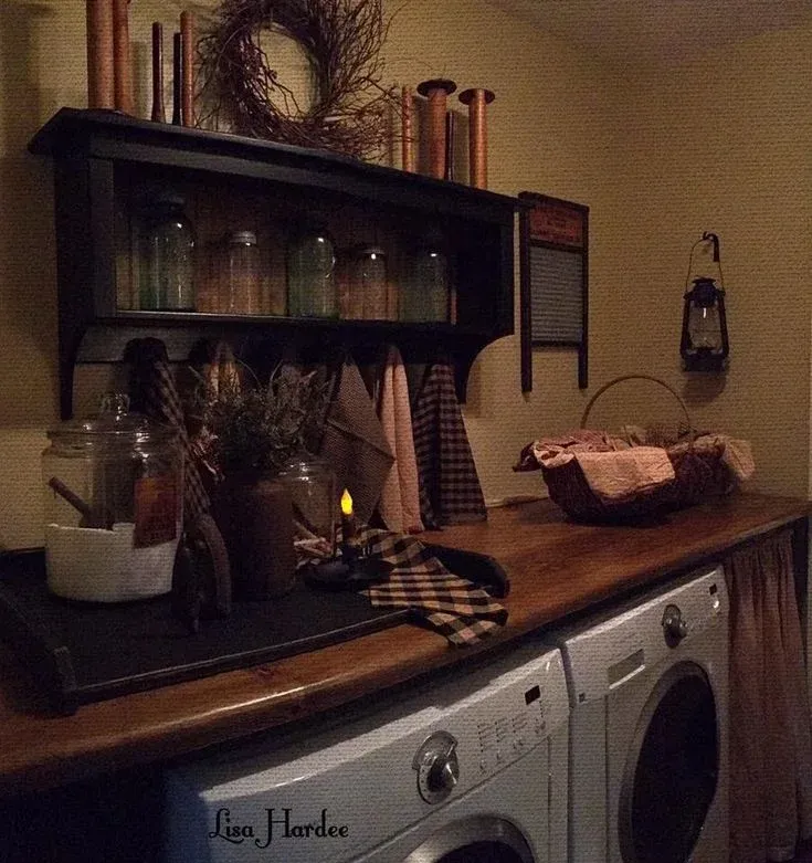 A warm, rustic country-style laundry room featuring a dark wooden shelf filled with mason jars and spools above a wooden folding counter, decorated with a twig wreath, plaid dish towels, glass canisters, a wall-mounted lantern, and a vintage washboard above front-loading appliances.