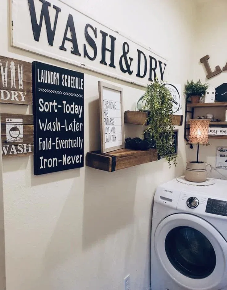 A decorated laundry room wall featuring wooden signs with texts like "WASH & DRY" and a humorous "LAUNDRY SCHEDULE" board, rustic floating shelves with greenery and framed art, and a white front-loading Samsung washing machine below.