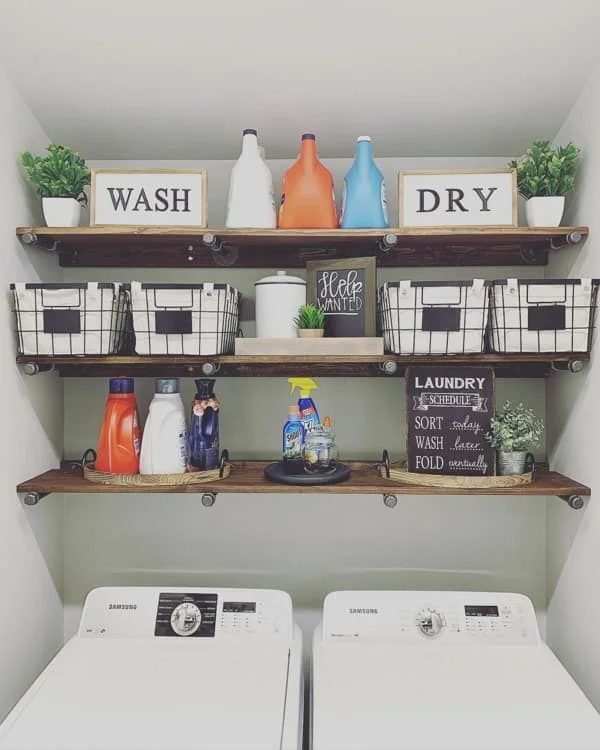 Three dark wood floating shelves mounted with black pipe brackets above a white Samsung washer and dryer nook. The shelves hold laundry detergents, black metal wire storage baskets with fabric liners, potted faux plants, and decorative framed signs reading "WASH" and "DRY".