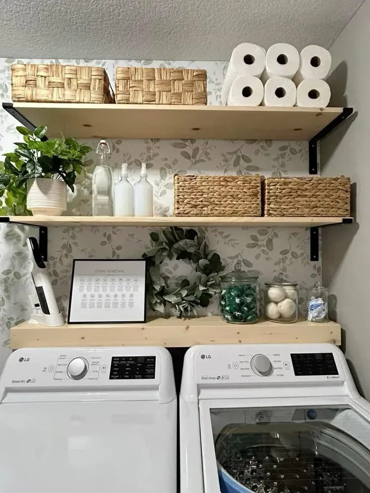 A stylish and practical laundry nook featuring two wall-mounted natural pine floating shelves with black metal hardware, plus a floating shelf bridge directly over modern LG appliances. The muted grey-and-white leaf wallpaper adds a subtle pattern behind neatly stored paper towels, woven baskets, clear dispenser bottles, and decorative greenery.