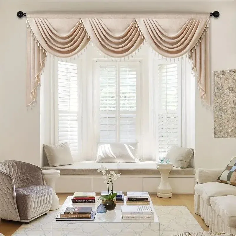 A formal living room window styled with a three-swag cream valance with tassel fringe, white interior shutters, and a built-in window seat.