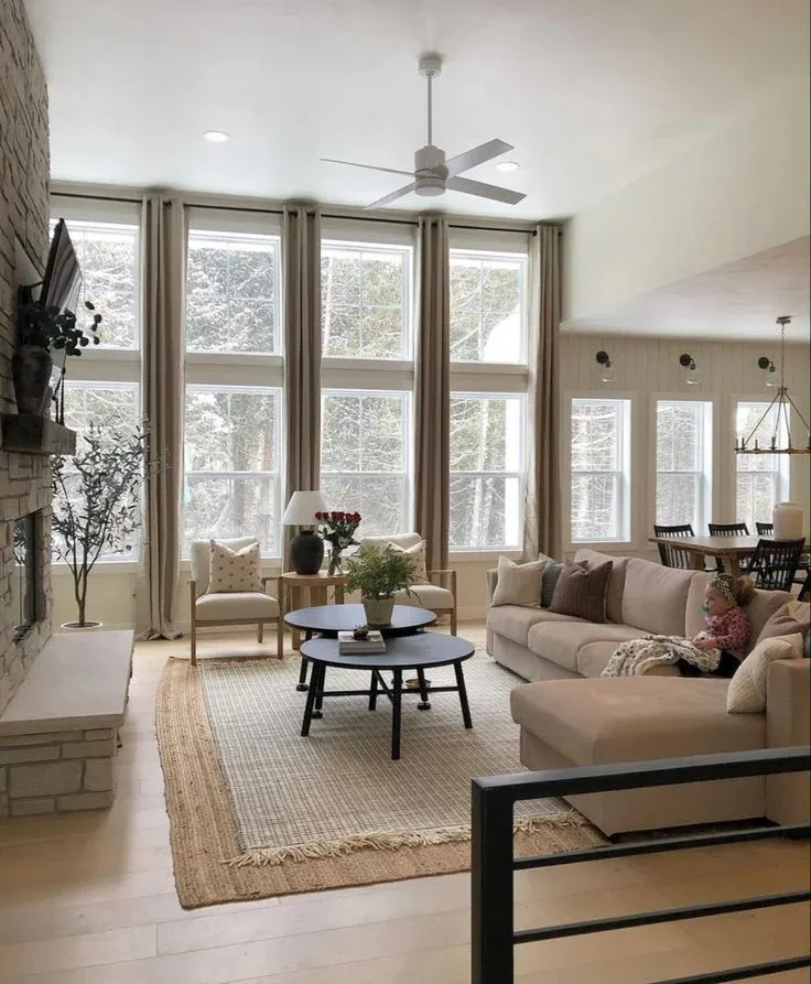 Bright living room featuring floor-to-ceiling windows styled with long beige curtains, a beige sectional sofa, stacked black round coffee tables, and a stone fireplace.