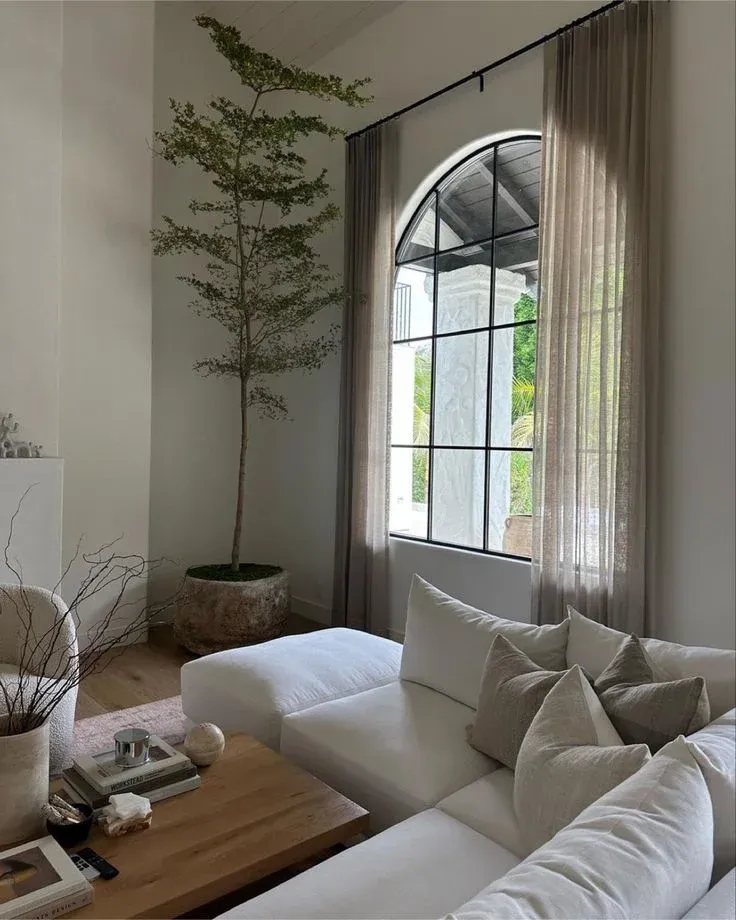 A tall black-framed arched window styled with floor-length sheer beige curtains in a modern living room, featuring a white sectional sofa and a large potted indoor tree.