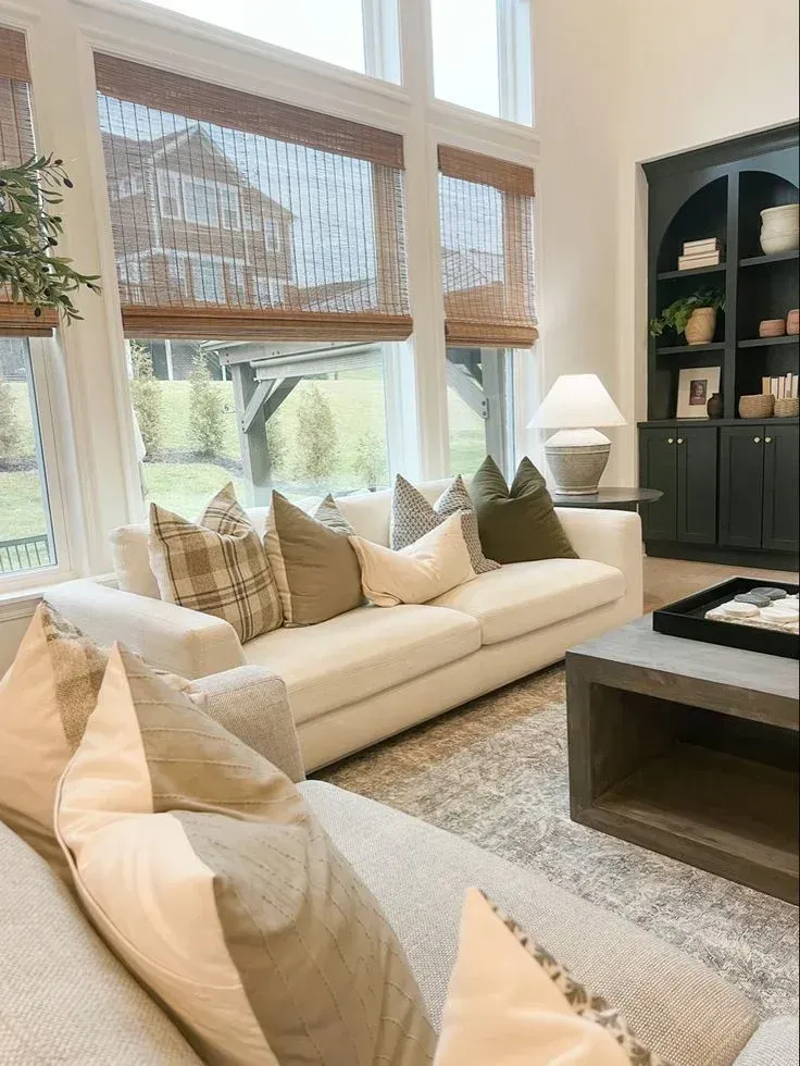Large living room windows styled with woven bamboo Roman shades, featuring a cream sofa adorned with plaid and olive green pillows, and a dark built-in bookcase.