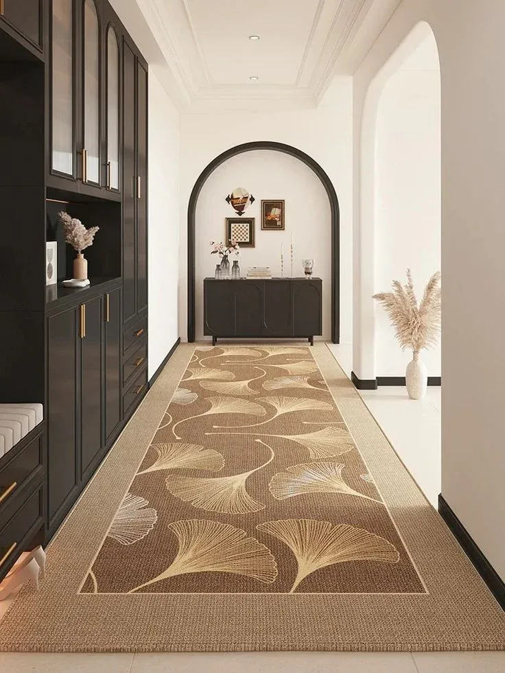 A spacious, elegant hallway featuring a large brown and gold ginkgo leaf patterned runner rug stretching down the center. The left wall is lined with seamless, built-in matte black cabinetry featuring gold hardware, while a grand black arched trim frames a view of a matching black credenza and curated artwork at the far end of the bright corridor.