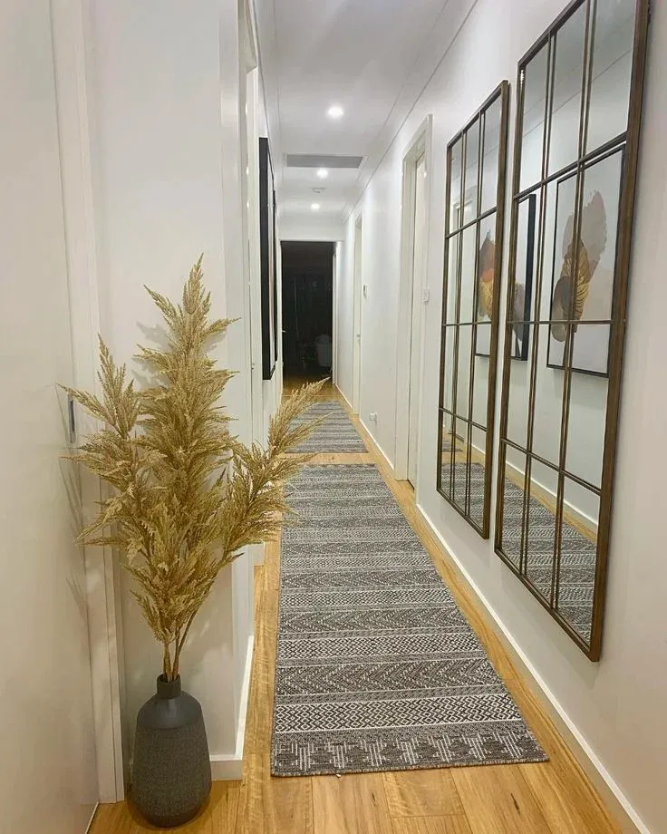 A long, narrow white hallway with light wood floors lined with a succession of grey and white geometric patterned runner rugs. The right wall features two large, vertical metal windowpane-style mirrors that reflect the opposing wall and framed artwork. In the left foreground, a tall black ceramic vase holds fluffy, dried pampas grass branches under bright recessed ceiling lights.
