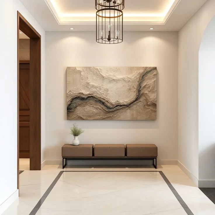Modern Foyer with Textured Canvas Art and Upholstered Bench A bright, modern hallway entry featuring white walls, polished marble flooring with a dark border, and a tray ceiling with a black cage pendant light. Centered on the main wall is a large, textured abstract painting in earth tones hanging above a long brown three-seater bench. A small white vase with greenery sits on the bench, flanked by a wooden doorway on the left and an arched opening on the right.