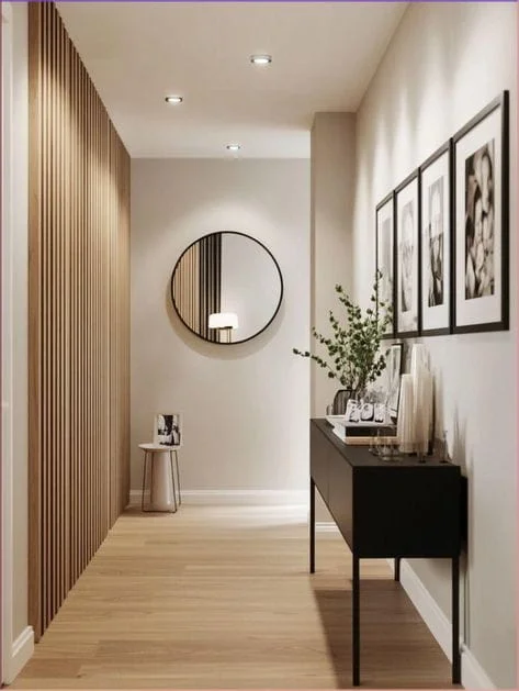 Modern Minimalist Hallway Interior Design A modern, minimalist hallway featuring a light wood slat accent wall on the left, a black console table decorated with art and greenery on the right, and a large round mirror mounted on the far wall above light wood flooring.