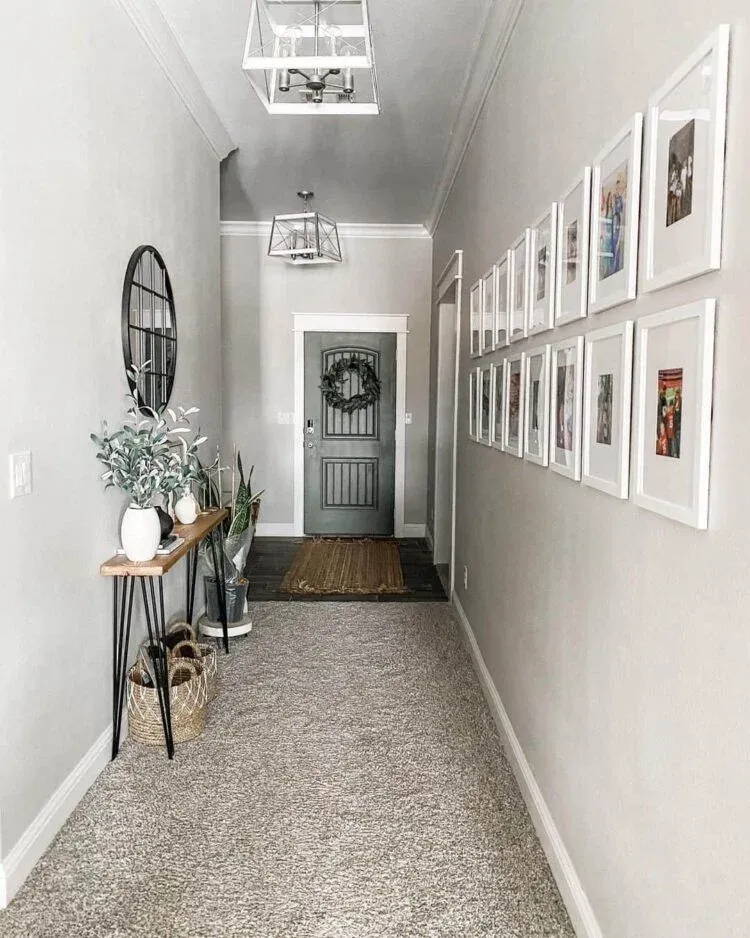Bright and airy narrow hallway decor showcasing a carpeted floor, lantern-style ceiling lights, a minimalist wooden console table, and a structured photo wall layout leading toward a stylish front door with a decorative green wreath.