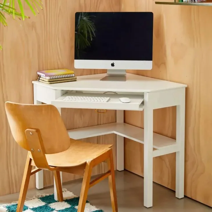Space-saving white wooden corner desk with an integrated keyboard slider holding a desktop computer, paired with a vintage-style wooden chair in a brightly lit room.